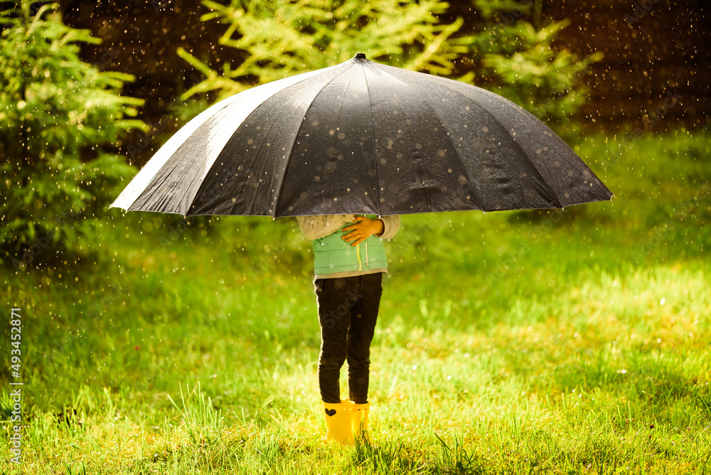 Little girl hid from spring or summer rain under huge silver umbrella. Girl dressed in trousers, jacket, yellow rubber boots.