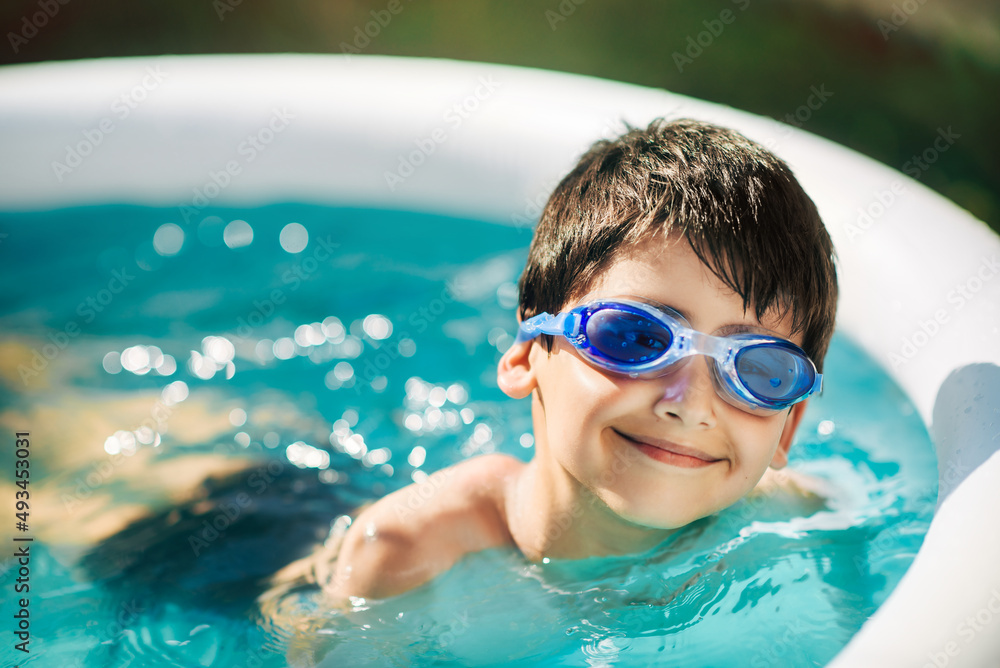Naklejka premium Little boy in swimming goggles is resting on edge of an inflatable circular pool outside on sunny summer day. Child is cheerful and happy. Close up. Summer vacation and healthy lifestyle concept.