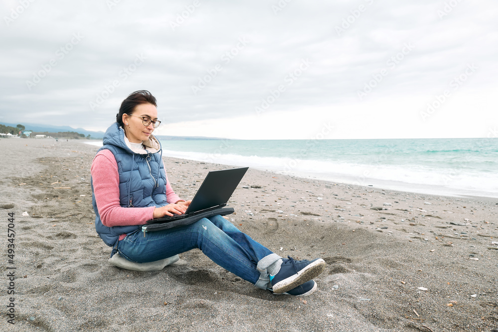 Beautiful sporty woman wearing eyeglasses with laptop computer working outdoors while sitting on winter beach in front of the sea. Modern lifestyle, connection, blogging, business, freelance work.