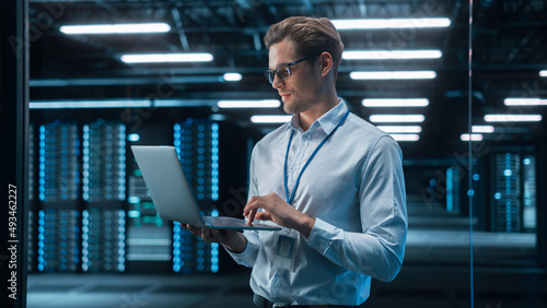 European Male IT Specialist Using Laptop Computer, Standing in Big Warehouse Data Center. System Administrator working with SAAS. E-Business Digital Entrepreneur Examining Cloud Web Services