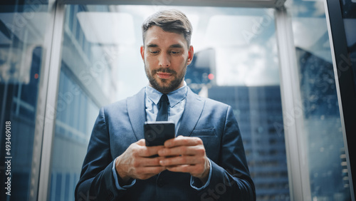Successful Businessman Riding Glass Elevator to Office in Modern Business Center. Handsome Happy Man Smile while Using Smartphone, Write Text Message, Check Social Media and Work Emails in a Lift.