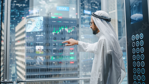 Successful Muslim Businessman in Traditional White Kandura Studying Stock Market Using Futuristic Holographic Screen in the Elevator. Male is Taking a Lift to the Office Inside the Buisness Center.