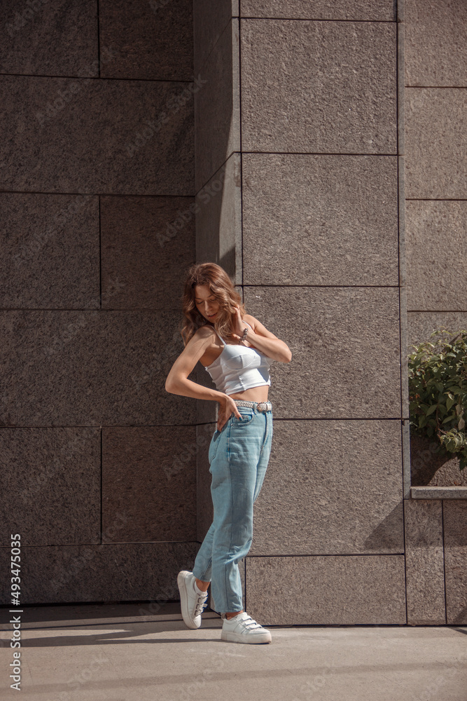 young curly brunette girl in white top and blue jeans is standing with flirt near store at sunny day and looking down with hand near hair. lifestyle concept, free space young curly brunette girl in white top and blue jeans is standing with flirt near store at sunny day and looking down with hand near hair. lifestyle concept, free space