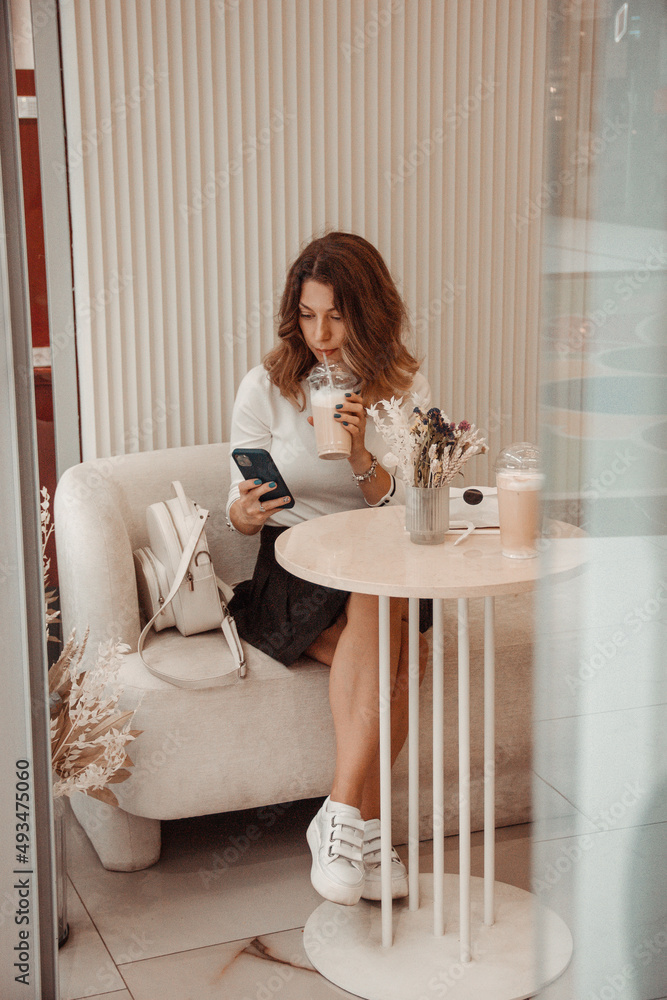 young curly brunette girl in white shirt and black skirt is sitting in beige modern cafe with mobile phone in hand and drinking ice coffee on the white wall background. lifestyle concept, free space