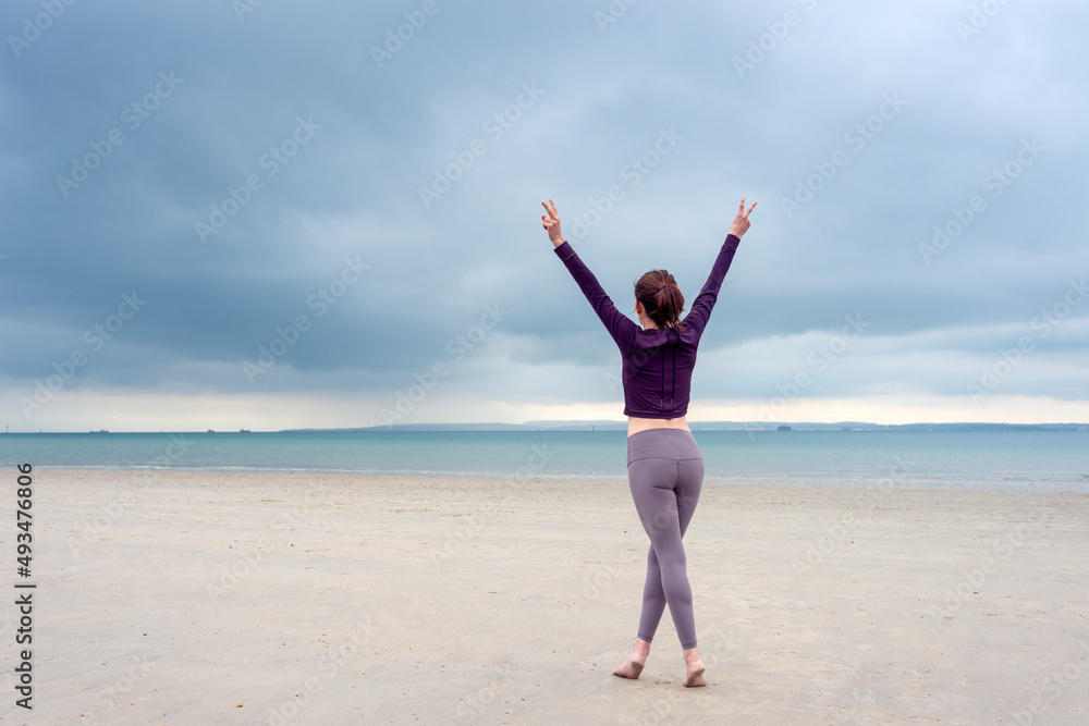 Young woman in sportswear is standing and lifting up arms for freedom on the beach, wearing fitnesswear