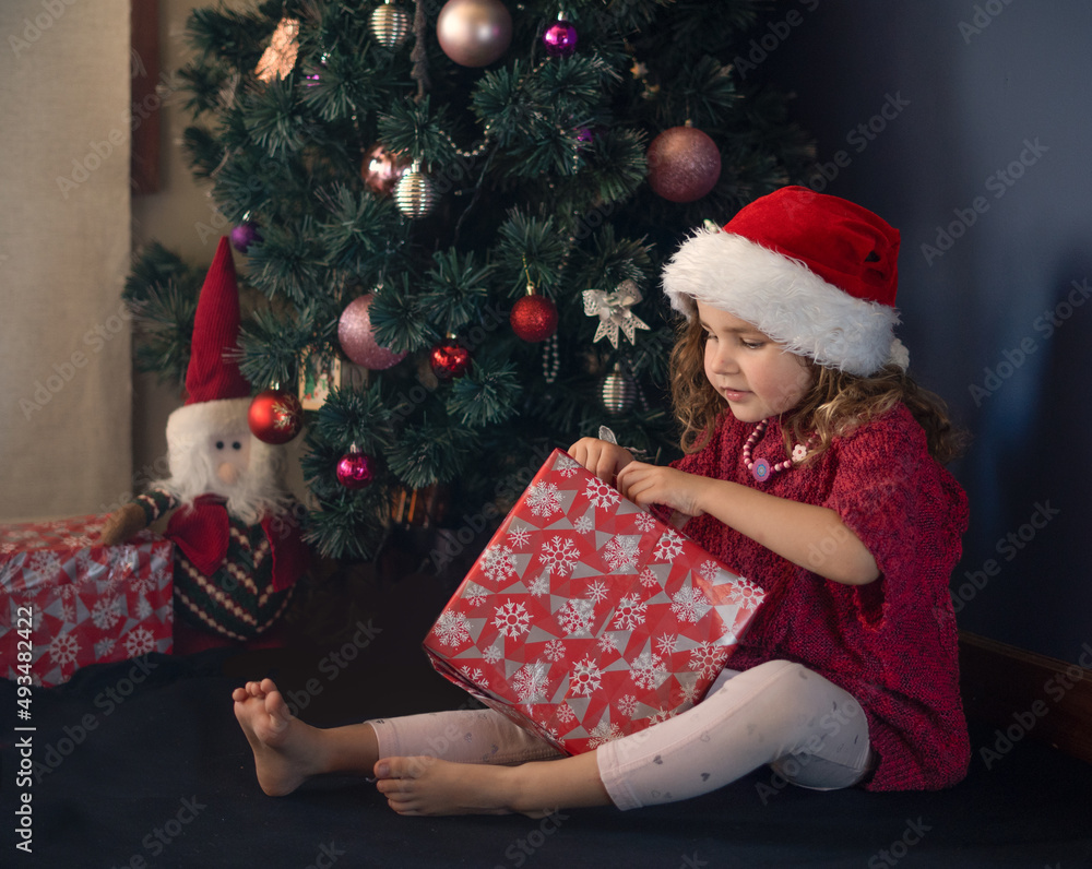 A little girl in red christmas hat and red sweater looking at her Christmas gift sitting near a Christmas tree. A little girl in red christmas hat and red sweater looking at her Christmas gift sitting near a Christmas tree.