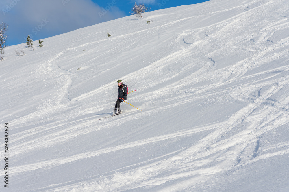 Woman with brown hair, wearing ski wear and a bagpack skiing down a beautiful snow hill with plenty of tracks on the snow, view from the side from far away, stryn norway, horizontal