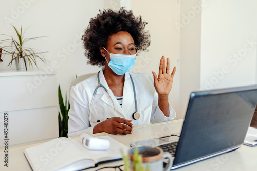 Female doctor wearing face mask, waving and talking with colleagues through a video call with a laptop in the consultation. Video conference, specialist talking, counseling, helping patient online.