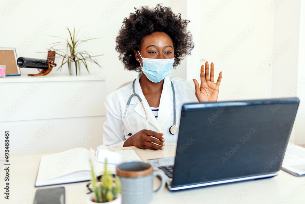 Female doctor wearing face mask, waving and talking with colleagues through a video call with a laptop in the consultation. Video conference, specialist talking, counseling, helping patient online.