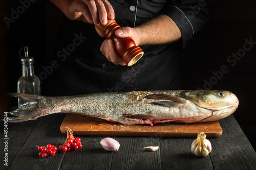 The chef prepares silver carp herring in the kitchen. The cook sprinkles pepper on the fish. Work environment on the kitchen table photo