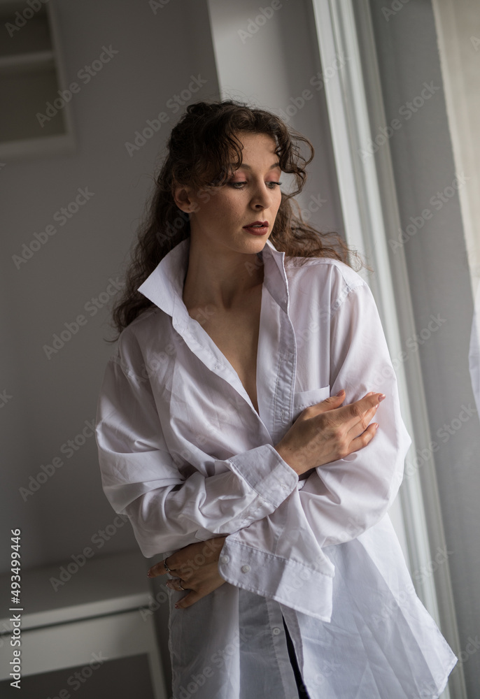 Attractive sexy brunette in white shirt posing provocatively, near a window,studio shot. Portrait of sensual woman with long hair, in classic boudoir scene , looked out the window Attractive sexy brunette in white shirt posing provocatively, near a window,studio shot. Portrait of sensual woman with long hair, in classic boudoir scene , looked out the window