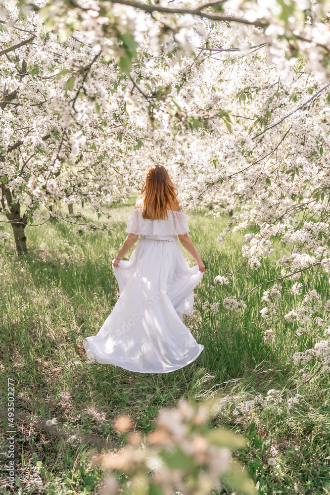 Fantasy woman in long white elegant fashion long dress walks in green spring blossom cherry garden. Happy cheerful girl princess bride. Skirt fabric flies flowing waving in wind motion.