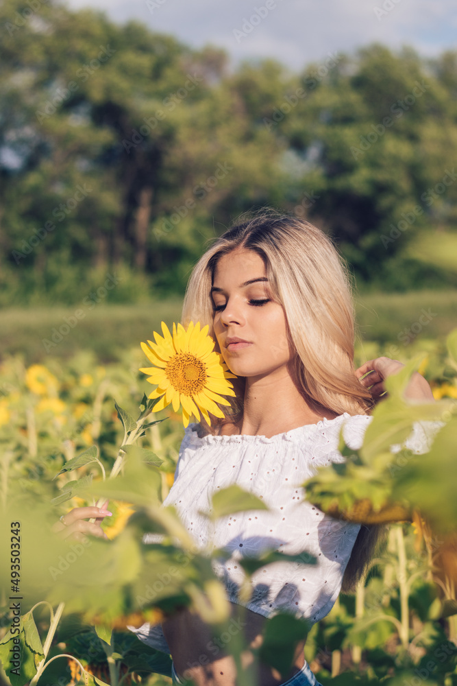 Beautiful young long-haired blonde woman in a white blouse with bare shoulders holds a sunflower in her hands on a sunflower field.Summer concept. Beautiful young long-haired blonde woman in a white blouse with bare shoulders holds a sunflower in her hands on a sunflower field.Summer concept.