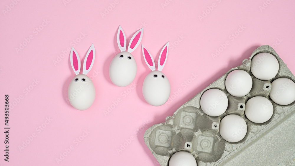 Card box with white natural eggs and eggs with rabbit ears on pastel pink background. Flat lay