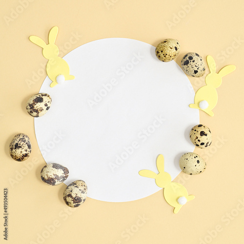 Round paper card copy space with yellow rabbits and natural quail eggs on pastel beige background. Flat lay Easter concept
