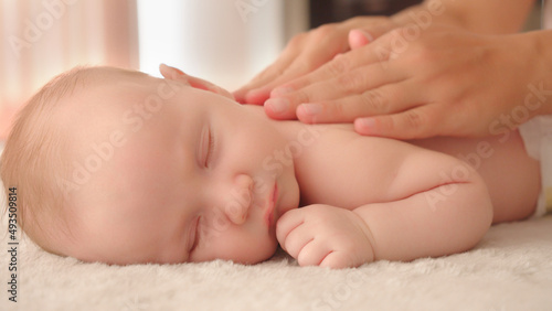 Mother doing massage on her healthy sleeping infant baby. Small caucasian newborn resting and laying on his belly while his mother is performing a massage for his small back and developing muscles. .