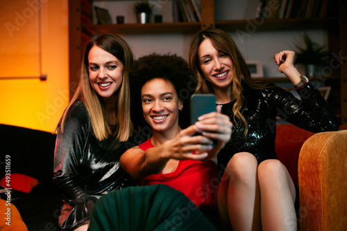 Three multicultural female friends sit in the living room in the evening and take selfies at home party.