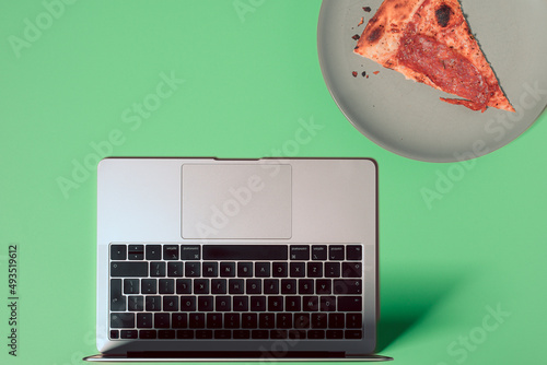 Overhead view of the colorful office desk with notebook laptop, and slice of pizza on plate on pastel color background
