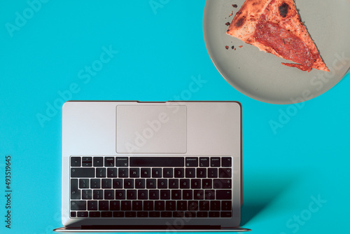 Overhead view of the colorful office desk with notebook laptop, and slice of pizza on plate on pastel color background
