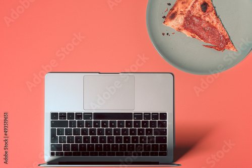 Overhead view of the colorful office desk with notebook laptop, and slice of pizza on plate on pastel color background
