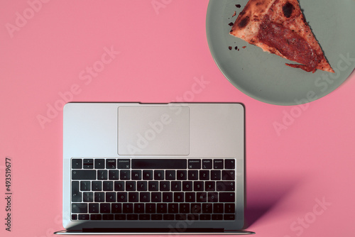 Overhead view of the colorful office desk with notebook laptop, and slice of pizza on plate on pastel color background
