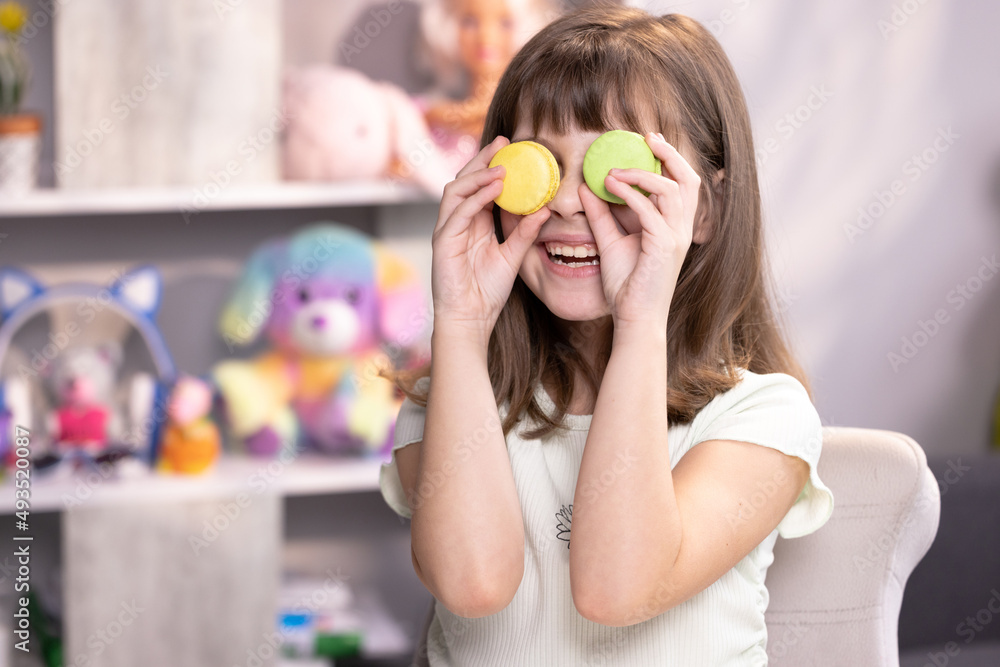 Happy smiling face of little girl covering her eyes with macaroons. School girl plays with dessert macarons, holding the cookies like glasses around the eyes and throws his hands to the sides Happy smiling face of little girl covering her eyes with macaroons. School girl plays with dessert macarons, holding the cookies like glasses around the eyes and throws his hands to the sides