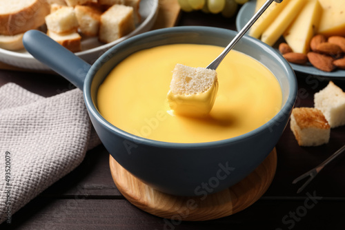 Dipping piece of bread into tasty cheese fondue at wooden table, closeup