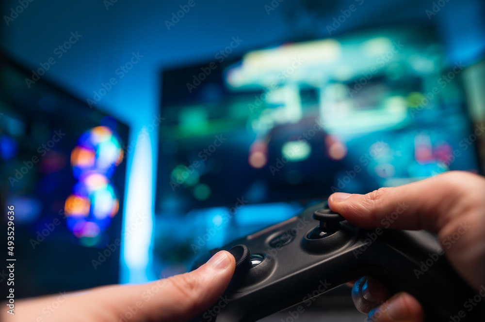 The gamer is playing a video game with friends. In his hands he has a modern joystick. Also in front is a large TV screen, personal computer monitor. Neon lighting. Video games with friends.