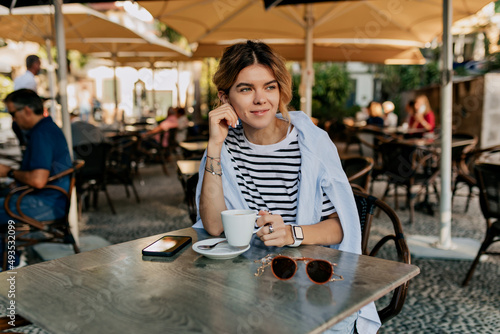 Stylish European woman with light hairstyle in striped t-shirt is resting on open air terrace with morning coffee. Elegant girl in white shirt relaxing with beverage in morning.