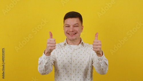 Happy smiling guy 20 years old in a white shirt shows thumbs up on a yellow background. OK photo