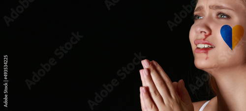 Scared praying woman with heart in colors of Ukrainian flag on her cheek against dark background. Stop the war