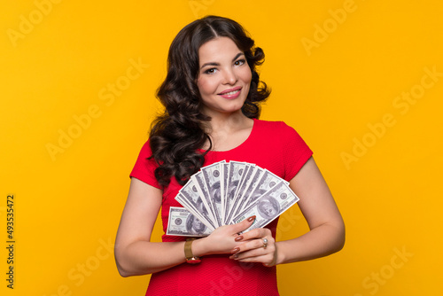Smiling businesswoman wears red dress showing fan of cash money in dollar banknotes in yellow background in studio. Rich girl looking at camera. Business, finance, saving, banking and people concept
