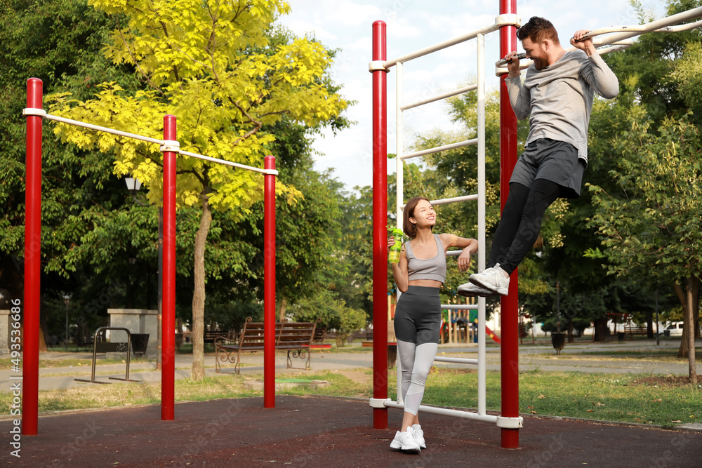 Fototapeta premium Young man doing chin-up with his wife on sport ground