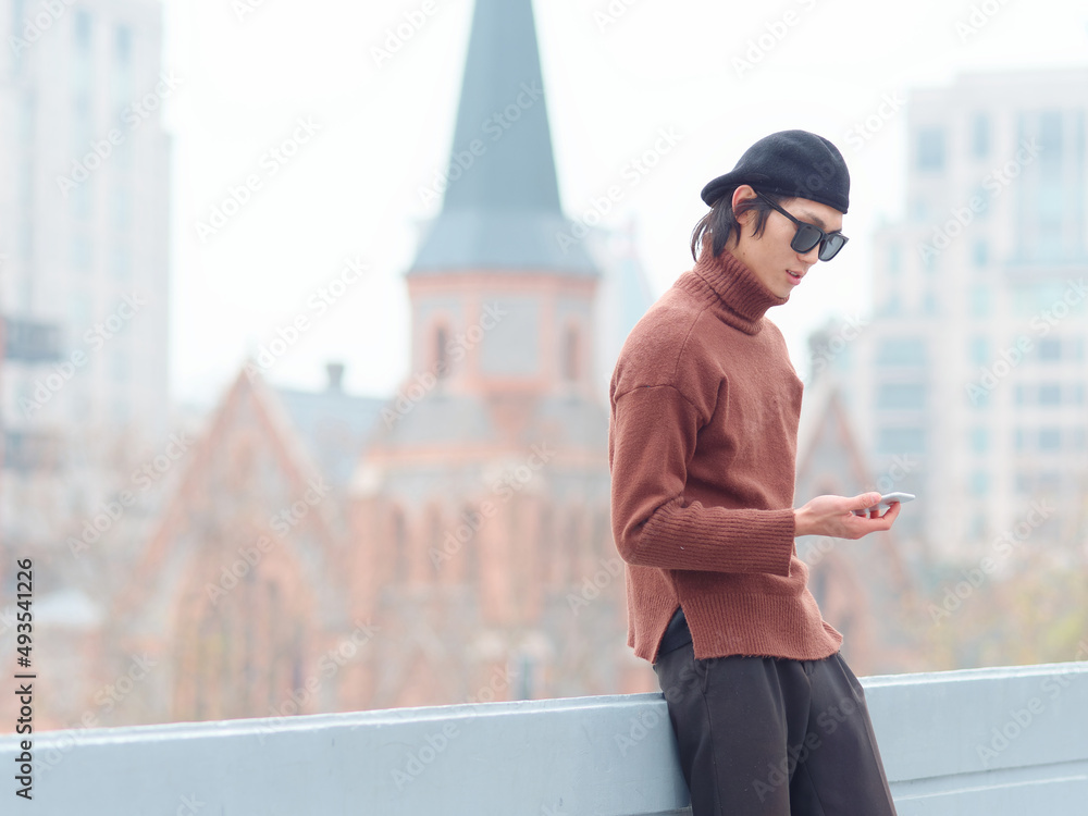 Portrait of handsome Chinese young man with sunglasses standing and looking at mobile phone with Shanghai city landmarks background, male fashion, cool Asian young man lifestyle.