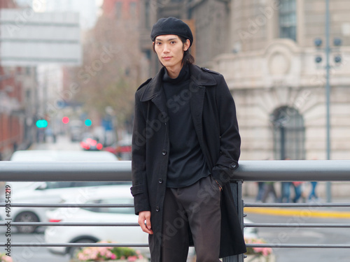 Portrait of handsome Chinese young man wearing black overcoat, sweater and trousers standing on Shanghai street, front view, male fashion, cool Asian young man lifestyle.