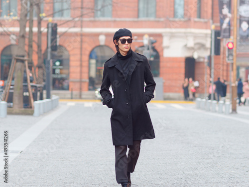 Portrait of handsome Chinese young man wearing black overcoat, sweater and trousers with sunglasses walking on Shanghai street, front view, male fashion, cool Asian young man lifestyle.