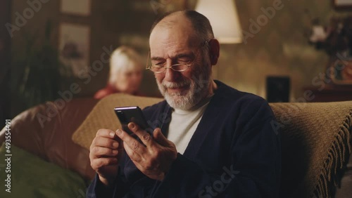 Pan around view of cheerful elderly male in glasses chuckling and reading news while sitting on sofa, and browsing social media on smartphone on weekend day at home