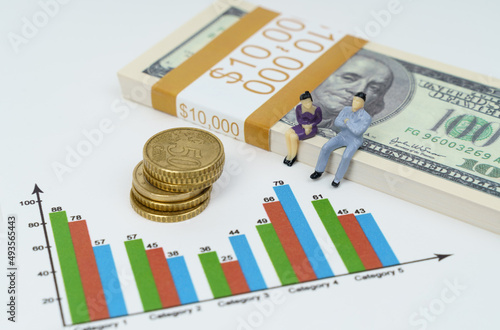 Miniature figures of businessmen sit on a pack of dollars. They look at the graph and talk. Coins next to them