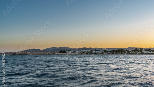 Evening in Egypt. A mountain range against the background of the sky, highlighted in orange. The houses of the hotel are visible far away on the shore. Ripples on the water. Copy space. Safaga
