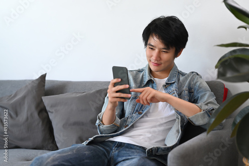 Happy man in casual wear using smart phone, checking social media, ordering delivery while relax on sofa.