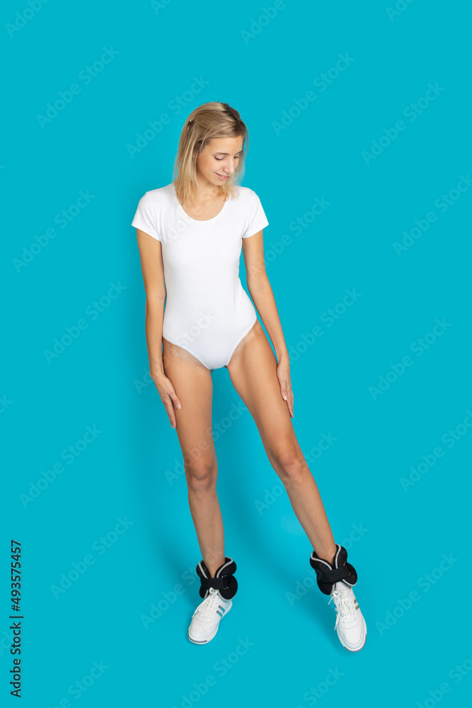 Exercising woman in white bodysuit and ankle weights looking to be in good shape posing on turquoise background. Healthy lifestyle, working out. Copy space. Exercising woman in white bodysuit and ankle weights looking to be in good shape posing on turquoise background. Healthy lifestyle, working out. Copy space.
