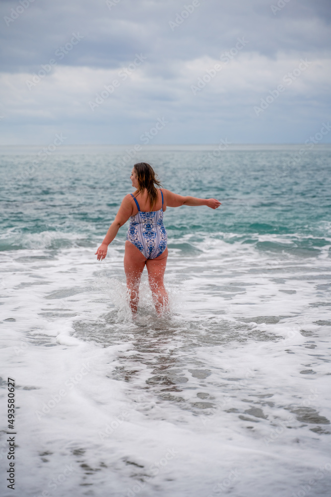 A plump woman in a bathing suit enters the water during the surf. Alone on the beach, Gray sky in the clouds, swimming in winter.