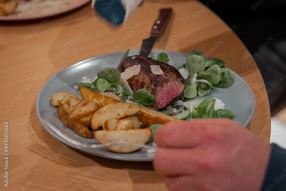 Hands are seen around a plate with juicy tender steak surrounded with vegetables and baked potatoes. Tasty food on a plate.