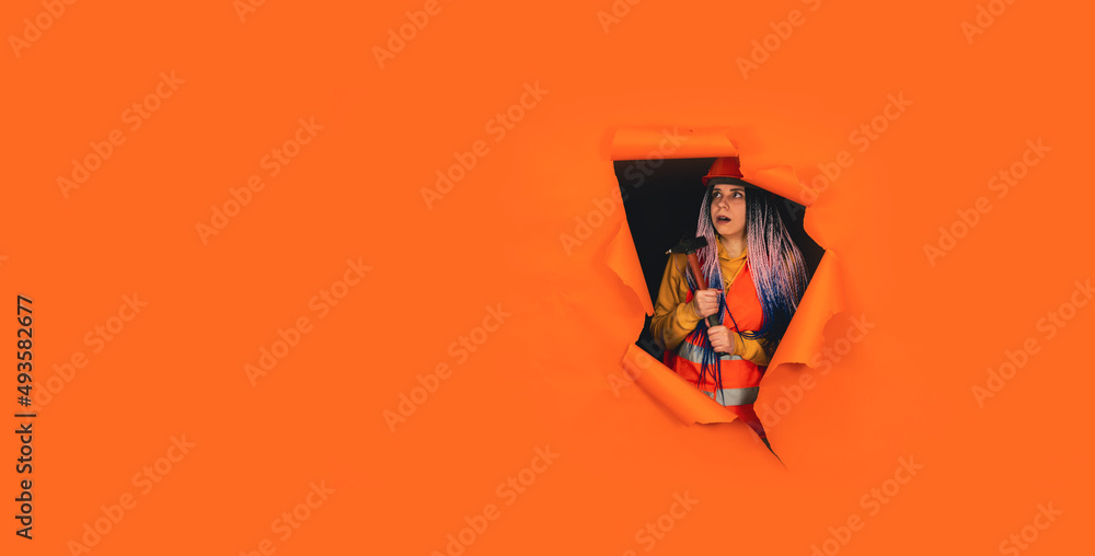 Shocked female construction worker in overalls with hammer looking out of hole of orange background. Young woman looking around in state of shock about what he did. Shocked female construction worker in overalls with hammer looking out of hole of orange background. Young woman looking around in state of shock about what he did.