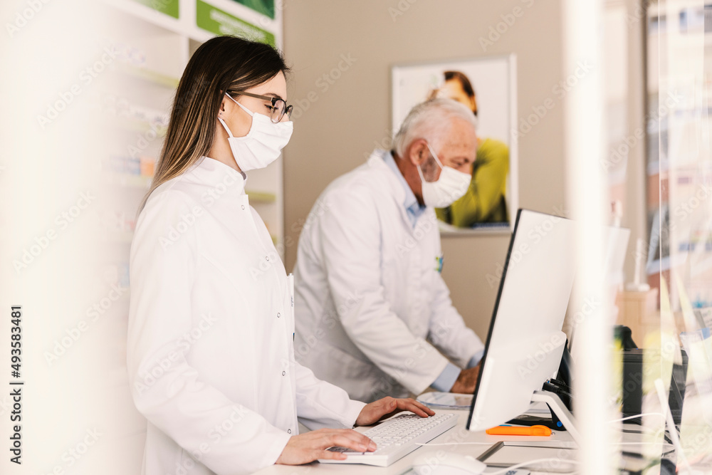Focused female pharmacist with mask using computer for data entry at pharmacy. Focused female pharmacist with mask using computer for data entry at pharmacy.