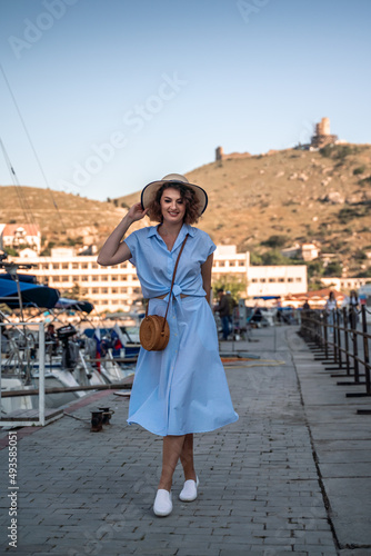 A young happy woman in a blue dress and hat stands near the seaport with luxury yachts. Travel and vacation concept © svetograph