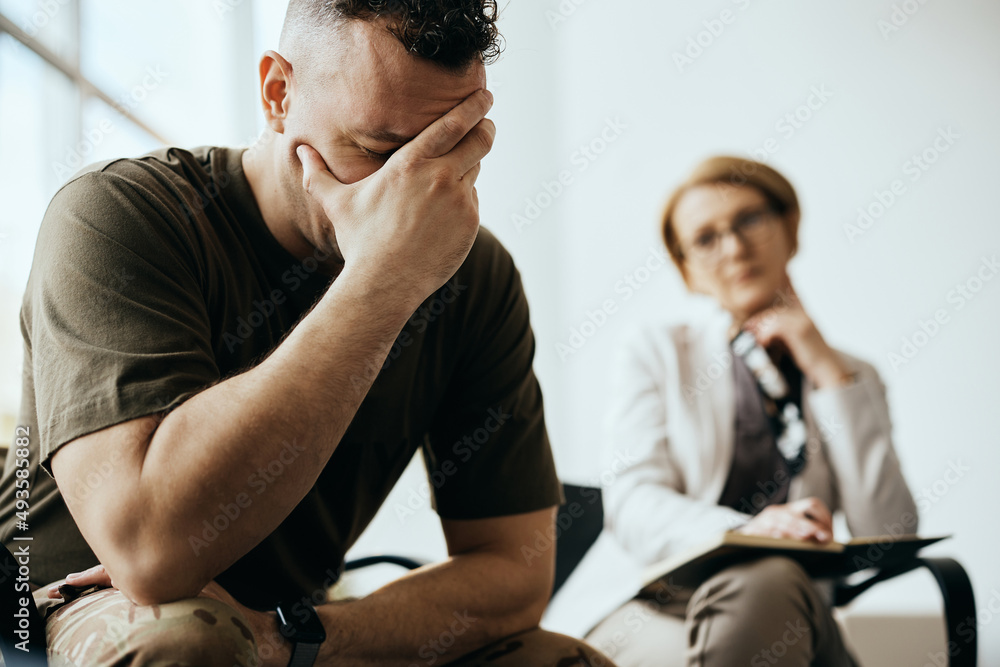 Stressed soldier holds his head in pain while talking about his issues with mental health professional. Stressed soldier holds his head in pain while talking about his issues with mental health professional.