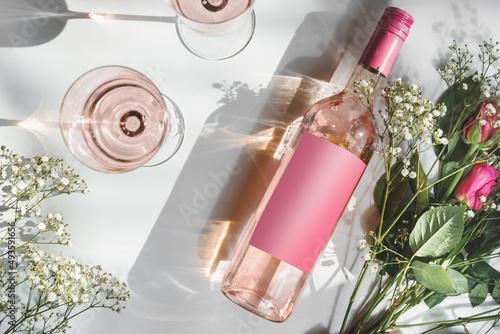 Rose wine bottle with empty pink mock up label, two wine glasses and romantic flower bouquet on white background with sunlight and shadow. Modern wine concept. Top view