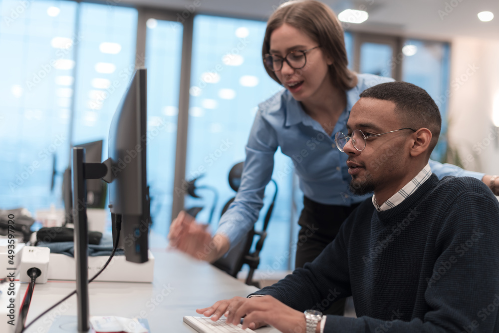Young smiling woman explaining to serious African American coworker project strategy. Diverse startup coworkers students woman and man talking discussing working in modern office using computer. Young smiling woman explaining to serious African American coworker project strategy. Diverse startup coworkers students woman and man talking discussing working in modern office using computer.