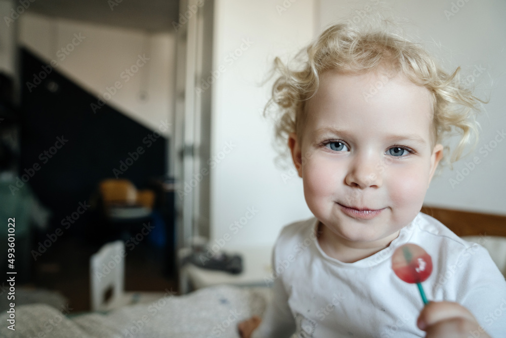 Fototapeta premium A cute baby looks into the camera and holds a sweet candy in his hand. Health and care for children's dental health.